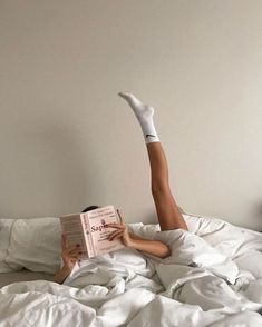 a woman laying in bed with her legs up on the pillow and reading a book