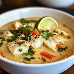 a white bowl filled with fish soup and garnished with cilantro, carrots, green herbs and lime