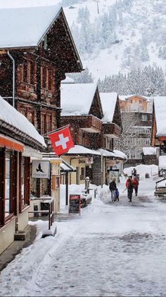 people walk down a snowy street lined with wooden buildings and flags in front of snow covered mountains