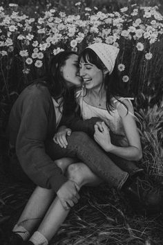 two women are sitting on the ground in front of some daisies and one is kissing her