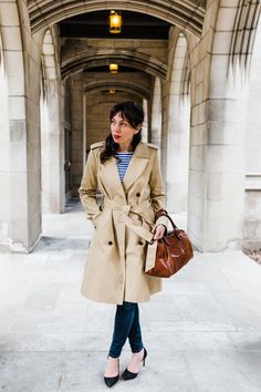 a woman in a trench coat is standing on the sidewalk holding a purse and looking off into the distance