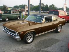 a brown muscle car parked in a parking lot next to other cars and people standing around