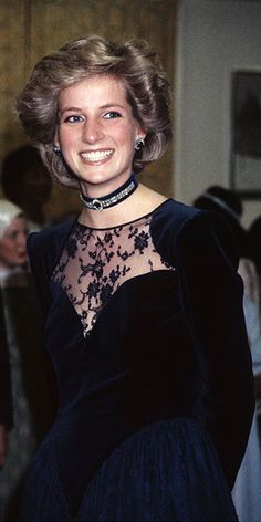 a woman in a black dress smiling and posing for the camera with other people behind her