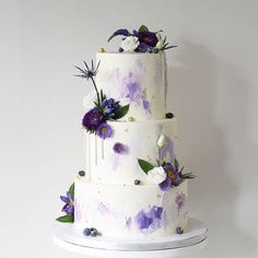 a three tiered white cake with purple flowers on the side and green leaves on top