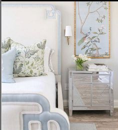 a white bed sitting next to a nightstand with flowers on it and a painting above it