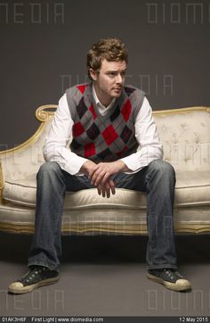 a man sitting on top of a white couch in front of a gray background wearing a red and black checkered vest