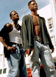 two young men standing next to each other in front of a white building with cars