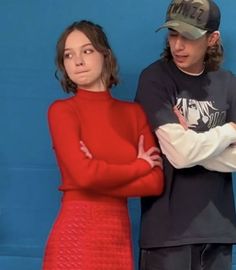 two people standing next to each other in front of a blue wall and one person wearing a baseball cap