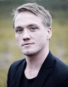 a close up of a person wearing a black suit and tie with blue eyes looking at the camera