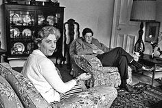 black and white photograph of two women sitting on couches in living room with china cabinet