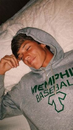 a young man laying in bed with his eyes closed wearing a grey hoodie that says michigan baseball