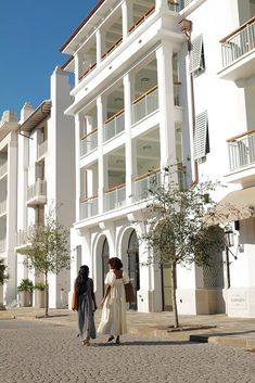 two women are walking down the street in front of some white buildings with balconies