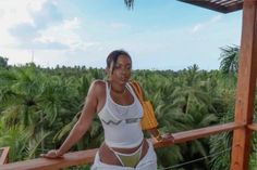 a woman standing on top of a wooden deck next to palm trees and holding onto a yellow purse