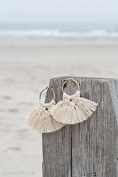 two tasseled earrings hanging from a wooden post on the beach with waves in the background