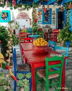 a red table surrounded by colorful chairs and tables with fruit on them in front of a blue building