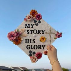 someone holding up a white graduation cap with flowers on it that says, my story his glory