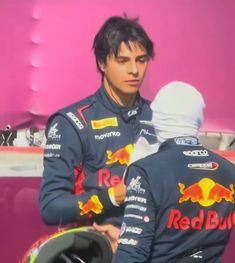 two men standing next to each other in front of a red bull car and one man holding a helmet