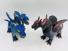 two toy dinosaurs are standing next to each other on a white surface, one is blue and the other is red