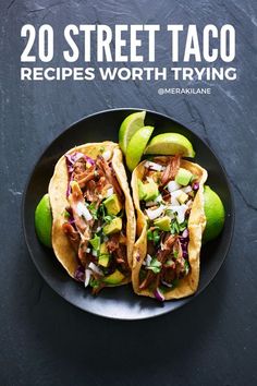 two tacos on a black plate with limes and avocado in the background