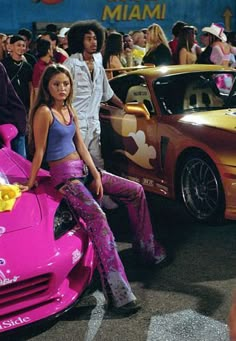 a woman sitting on the hood of a pink sports car in front of other people