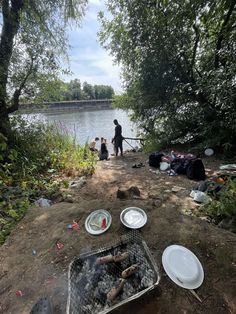 there are many plates and food on the ground by the water with people standing around