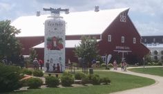 a large red barn sitting next to a lush green field with people walking around it