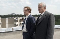 two men in suits standing next to each other on top of a roof with buildings in the background