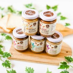 four jars of peanut butter sitting on top of a wooden cutting board next to green leaves