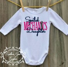 a white bodysuit with the words special mechanics daughter hanging from a clothes line on a wooden floor