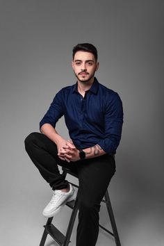 a man sitting on top of a stool wearing a blue shirt and black pants with his hands in his pockets