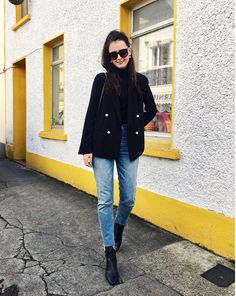 a woman standing in front of a yellow and white building wearing black jacket, jeans and boots