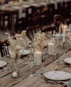 the table is set with white plates and silverware, candles and flowers in glass vases