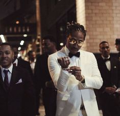 a man in a white tuxedo is looking at his cell phone while others stand around him