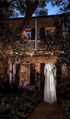 a white dress is hanging on a tree in front of a house with lights strung from it