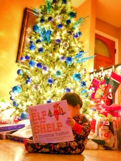 a young boy sitting in front of a christmas tree holding a sign that says elf on the shelf