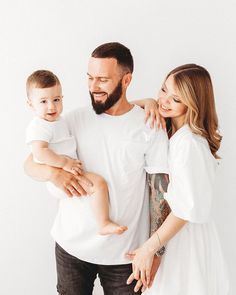 a man and woman holding a baby while standing next to each other in front of a white wall