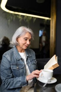 an older woman sitting at a table reading a book and holding a coffee cup in her hand