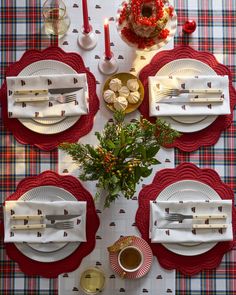 the table is set for christmas dinner with red and white plates, silverware, holly wreaths and candles