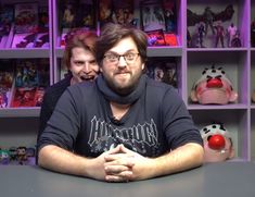 two people sitting at a table in front of bookshelves with stuffed animals on them