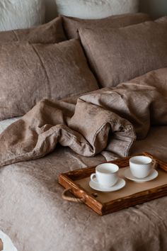 two white cups and saucers sitting on top of a bed with brown linens