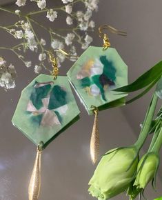 two green and gold earrings sitting on top of a table next to some white flowers