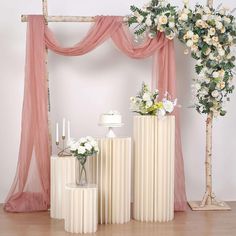 three white vases with flowers on them near a cake stand and flower arrangement in front of a pink drape