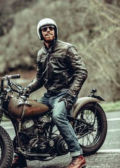 a man sitting on top of a motorcycle wearing a helmet and leather jacket with trees in the background