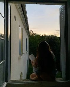 a woman sitting on a window sill looking out at the sunset and reading a book