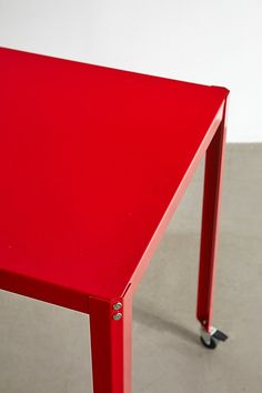 a red table sitting on top of a cement floor next to a white wall with wheels