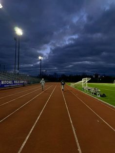 two men running on a track at night with the lights on and dark clouds in the background