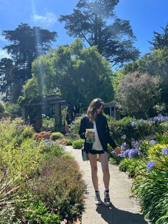 a woman walking down a sidewalk next to lots of flowers and trees in the background