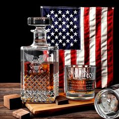 an eagle decanter and two shot glasses sit on a wooden cutting board with the american flag in the background