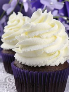 two cupcakes with white frosting sitting on a doily next to purple flowers