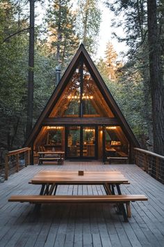 a frame cabin with picnic tables and lights on the porch in front of it, surrounded by tall trees